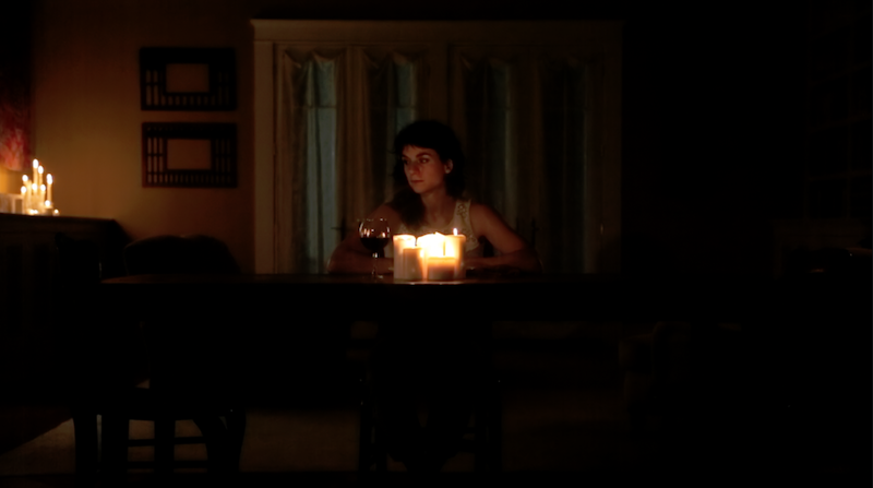Molly Lieber who plays Jeanine stands behind a piano. A glass of red wine sits besides her. The room is dimly lit by candelight. 
